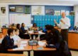 A United Charter School's teacher stands at the front of a classroom speaking to students seated in groups at tables, with blue lockers and educational posters in the background.