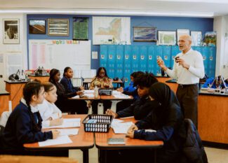 A United Charter School's teacher stands at the front of a classroom speaking to students seated in groups at tables, with blue lockers and educational posters in the background.