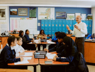 A United Charter School's teacher stands at the front of a classroom speaking to students seated in groups at tables, with blue lockers and educational posters in the background.