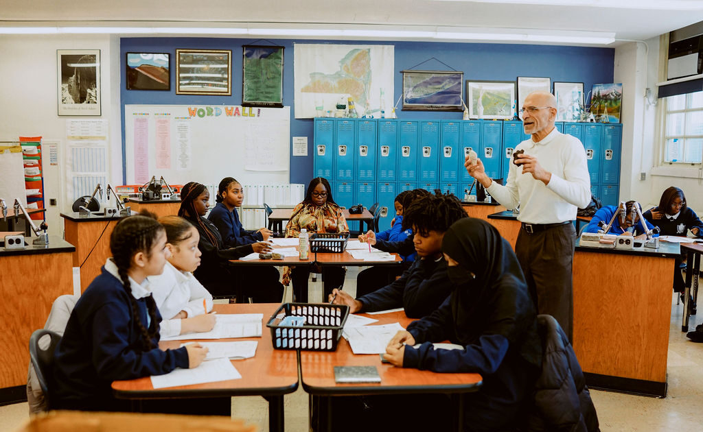 A United Charter School's teacher stands at the front of a classroom speaking to students seated in groups at tables, with blue lockers and educational posters in the background.