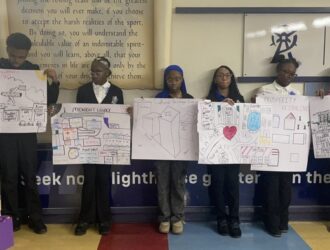 Seven students stand in a row indoors, each holding a large poster with hand-drawn diagrams and illustrations, likely presenting a group project.