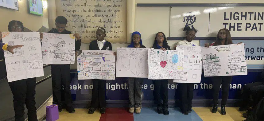 Seven students stand in a row indoors, each holding a large poster with hand-drawn diagrams and illustrations, likely presenting a group project.