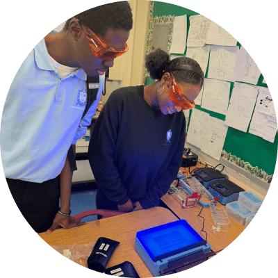 Two students wearing safety glasses observe a blue-lit electrophoresis gel in a classroom lab setting, with science equipment and diagrams visible in the background.