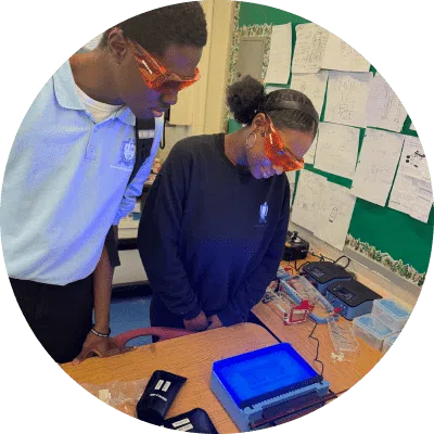 Two students wearing safety glasses observe a blue-lit electrophoresis gel in a classroom lab setting, with science equipment and diagrams visible in the background.