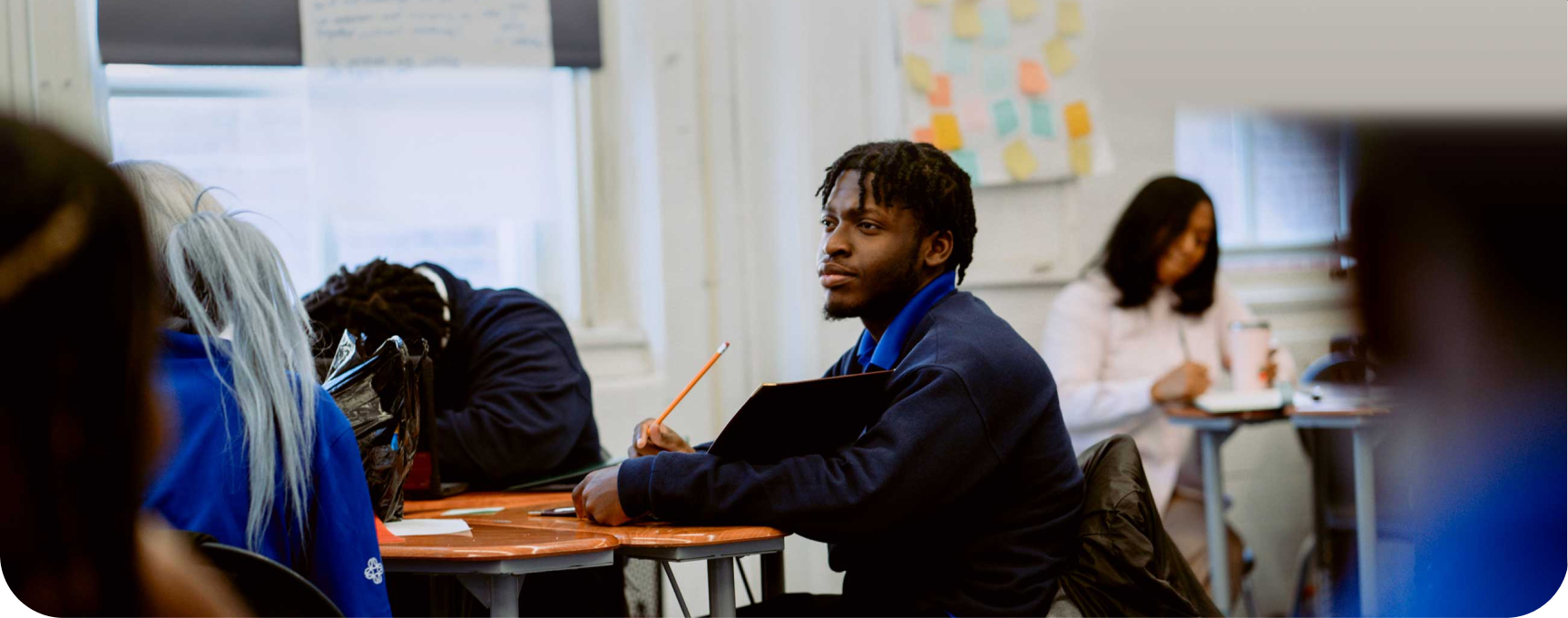 A student sits at his desk holding a pencil and notebook in a classroom, with other students and a teacher visible in the background.