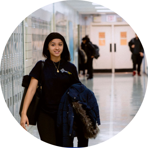 A student walks down a school hallway holding a coat and a bag, with lockers on one side and other people in the background.