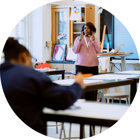 A teacher stands at the front of a classroom speaking, while a student sits at a desk in the foreground, facing the teacher.