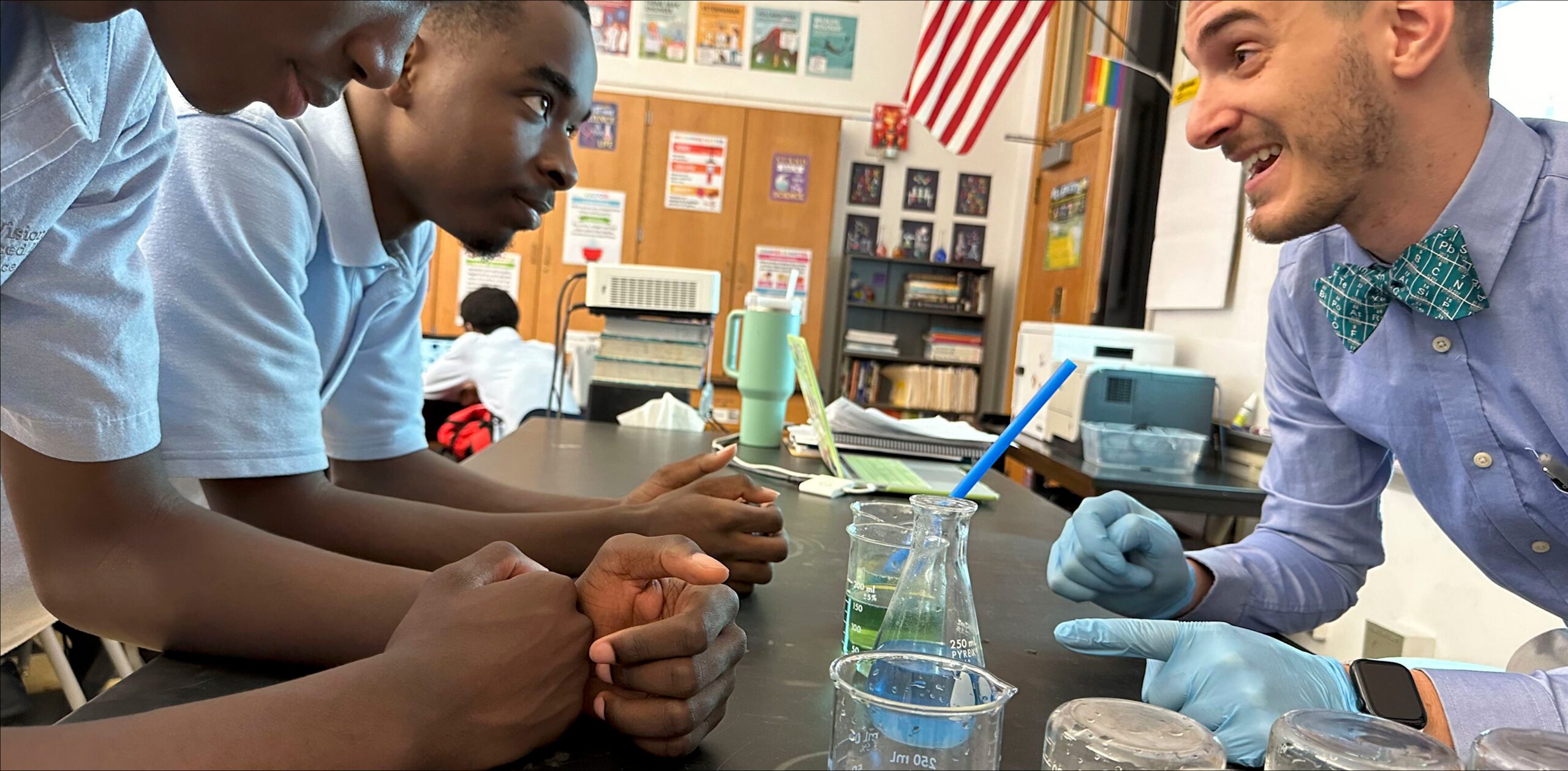 Three students listen to a teacher in a classroom during a science experiment, with beakers and lab equipment on the table.