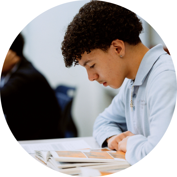 A young person with curly hair reads a book at a table, focused on the pages in front of them.