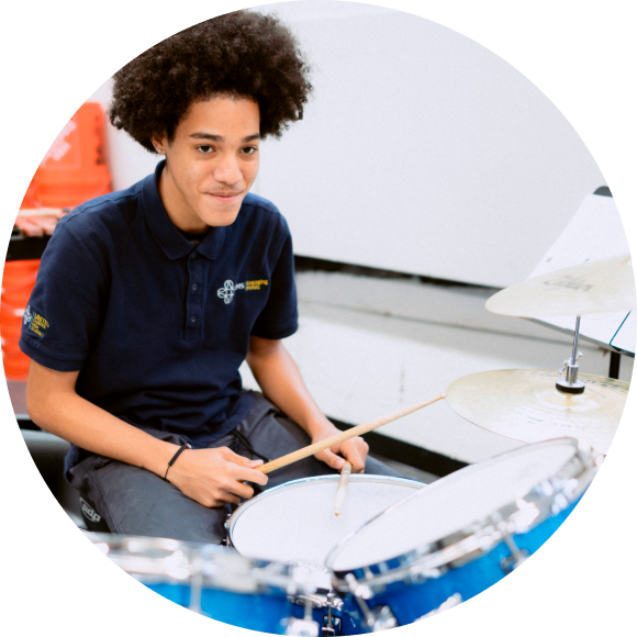 A young person in a navy polo shirt sits at a blue drum set, holding drumsticks and looking slightly upward, with a neutral white background.