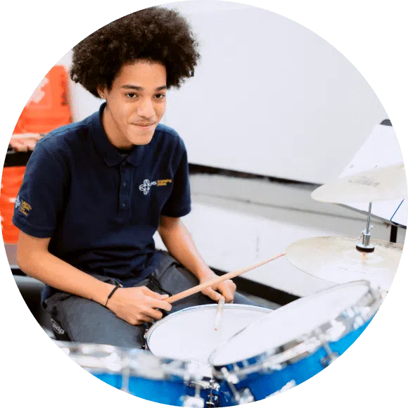 A young person in a navy polo shirt sits at a blue drum set, holding drumsticks and looking slightly upward, with a neutral white background.