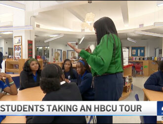 A group of NYC students sit at tables in a school library, listening to a standing adult woman during an HBCU tour event.