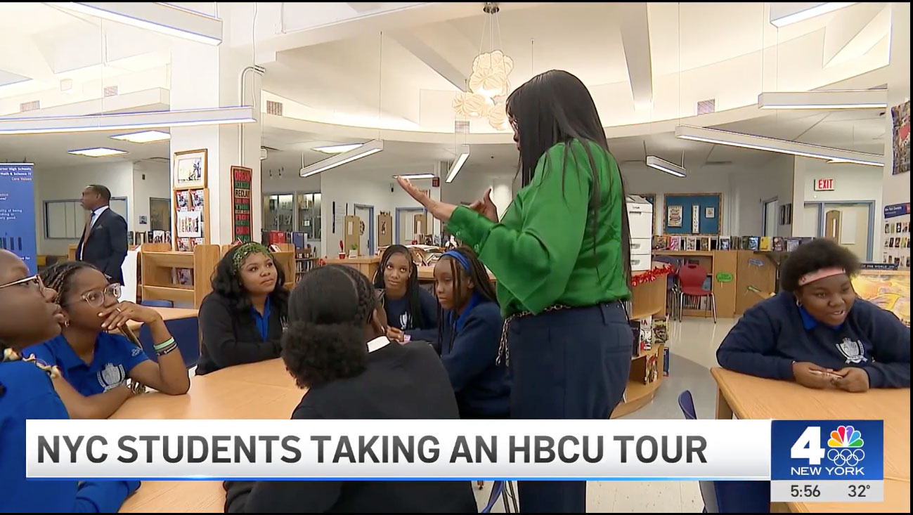A group of NYC students sit at tables in a school library, listening to a standing adult woman during an HBCU tour event.