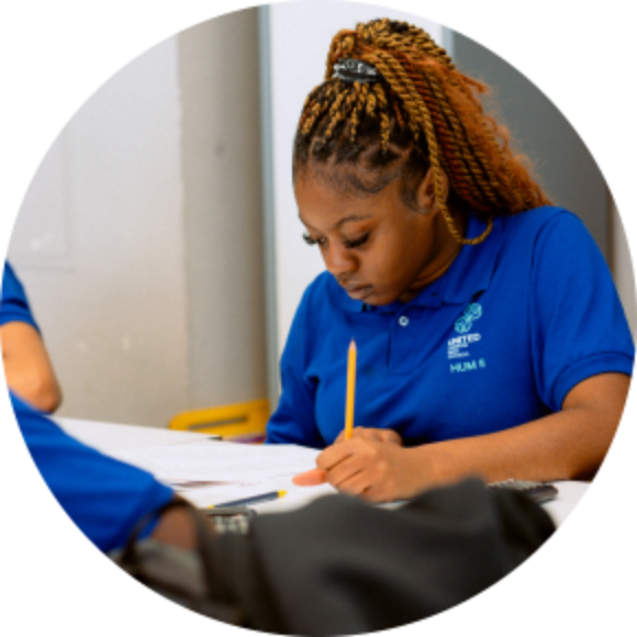 A young woman in a blue polo shirt writes on paper at a table, holding a pencil and concentrating on her work.