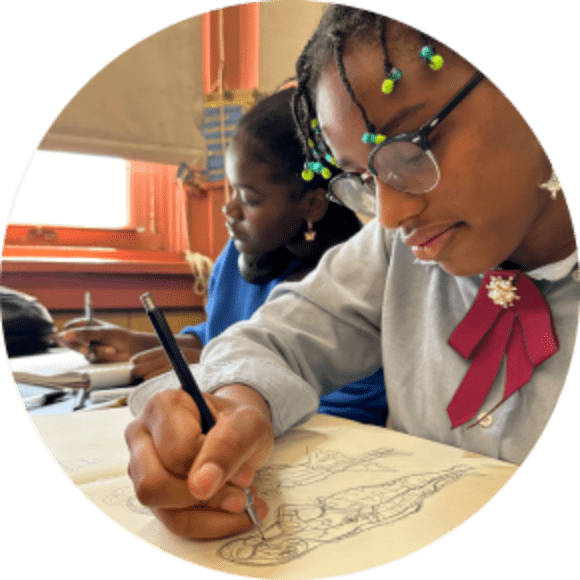 Two students sit at a desk in a classroom, focusing on drawing and writing in notebooks.
