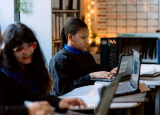 Three students sit at desks using laptops in a classroom. One student focuses on her screen, while the others are slightly out of focus in the foreground.