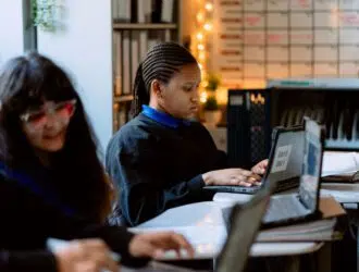 Three students sit at desks using laptops in a classroom. One student focuses on her screen, while the others are slightly out of focus in the foreground.