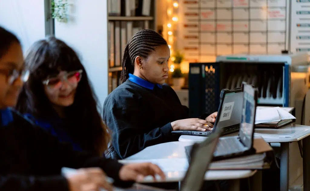 Three students sit at desks using laptops in a classroom. One student focuses on her screen, while the others are slightly out of focus in the foreground.