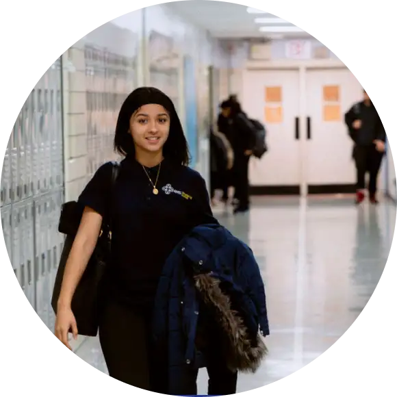 A student walks down a school hallway holding a coat, with lockers on the left and other people visible in the background.