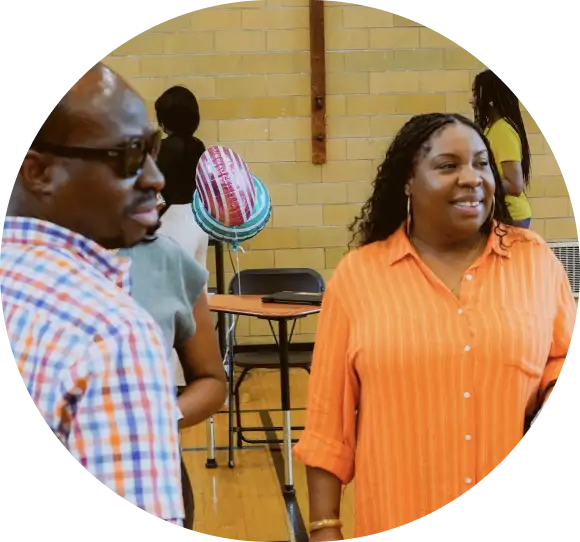 Two people standing and talking in a gymnasium, with a table, chairs, and balloon decorations in the background.