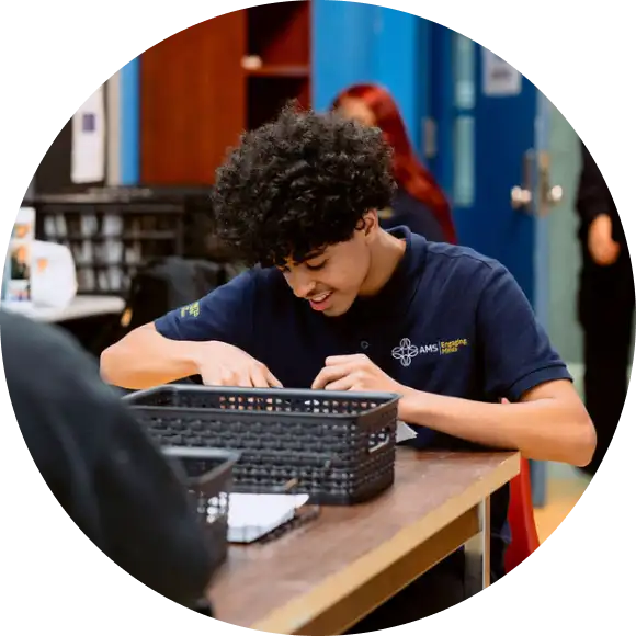 A student in a navy uniform shirt sits at a table and works on an activity with a black basket in front of him in a classroom setting.