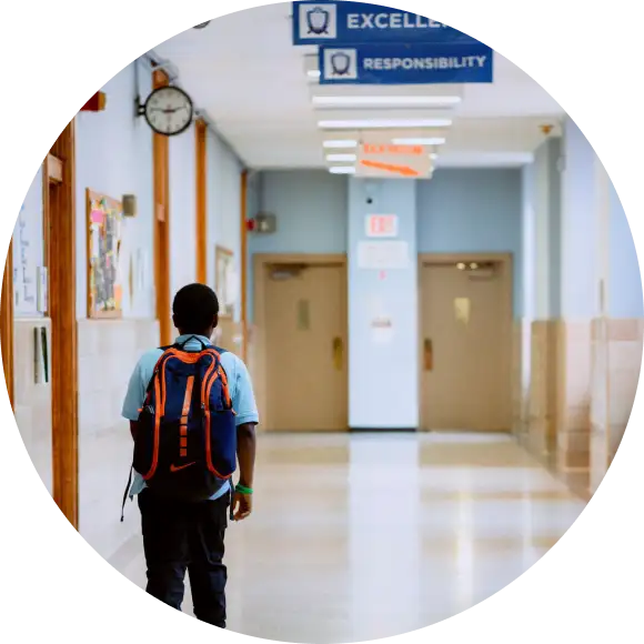 A student with a backpack walks alone down a school hallway with signs reading "Excellence" and "Responsibility" hanging from the ceiling.