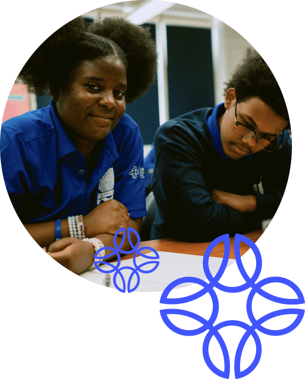 Two students in blue uniforms sit at a table; one is smiling at the camera while the other looks down at their work. Blue geometric graphics overlay the image.