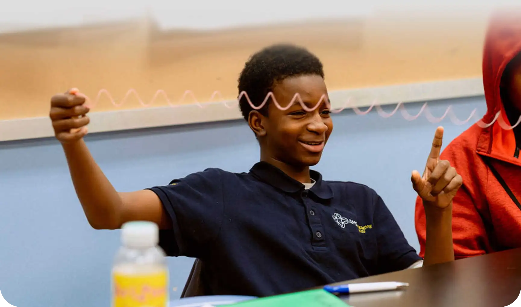 A student sits at a table, smiling and holding a spring toy stretched between his hands. A water bottle, folder, and pen are on the table in front of him.