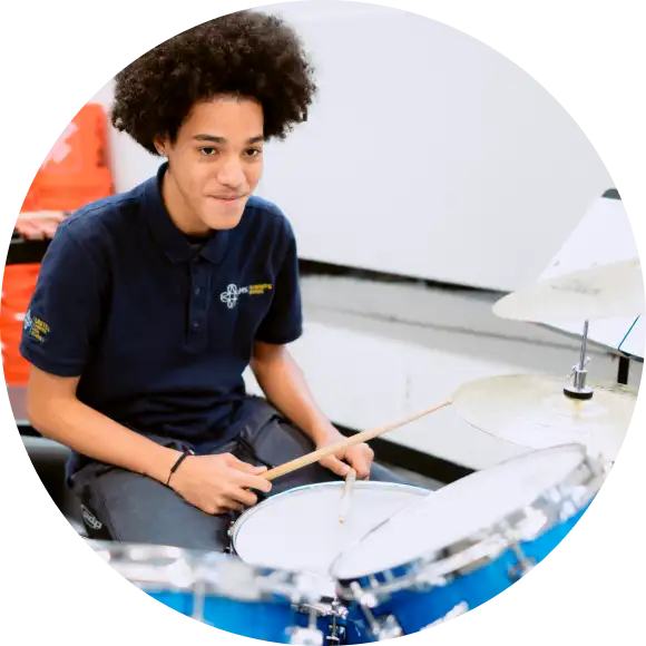 A person with curly hair plays a drum set indoors, holding drumsticks and wearing a navy blue polo shirt.