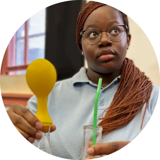 A woman wearing glasses holds a yellow balloon over a bottle with one hand and a green straw in a glass with the other.