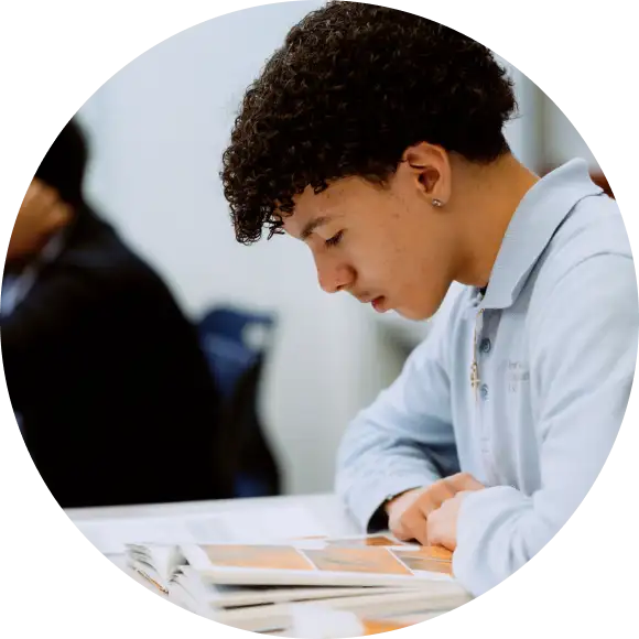 A young person with curly hair reads a book at a table, wearing a light blue polo shirt. Another person is blurred in the background.