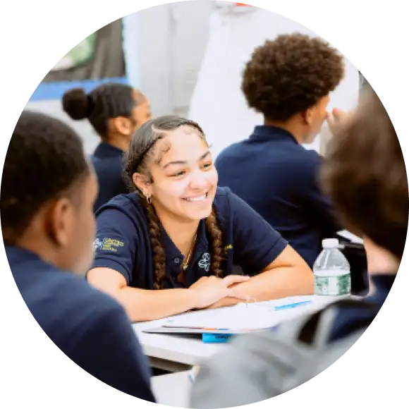 A student with braided hair smiles while sitting at a table with classmates in a classroom setting.