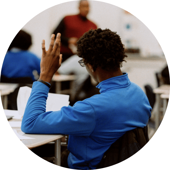 A student in a blue shirt sits at a desk and raises their hand in a classroom setting, with a teacher standing in the background.