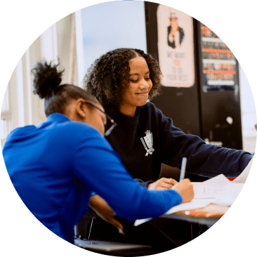 Two students sit at a desk working on papers, one writing while the other points at a document, in a classroom setting.