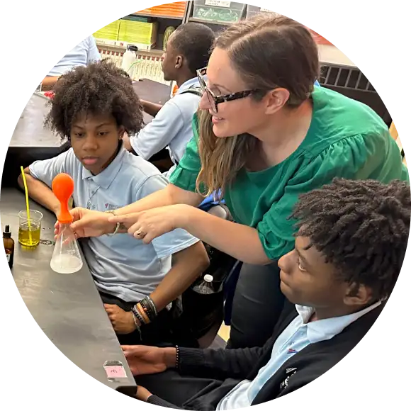 A teacher demonstrates a science experiment with a balloon on a flask to two students seated at a lab table.