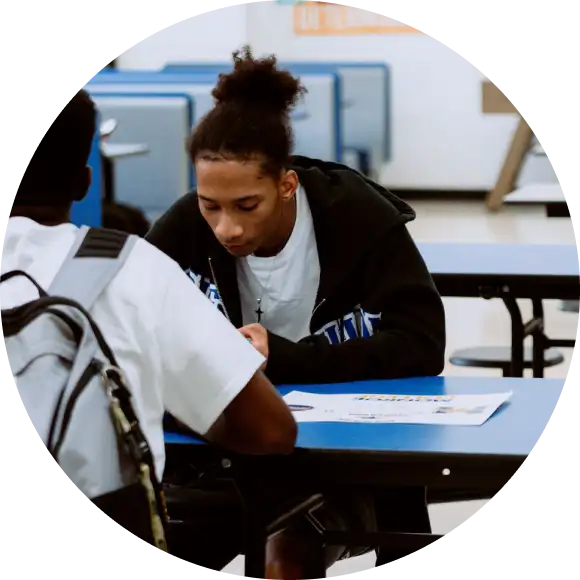 Two students sit at a blue table in a classroom or cafeteria, focused on papers in front of them. One wears a backpack, and the background shows blue booths and white walls.