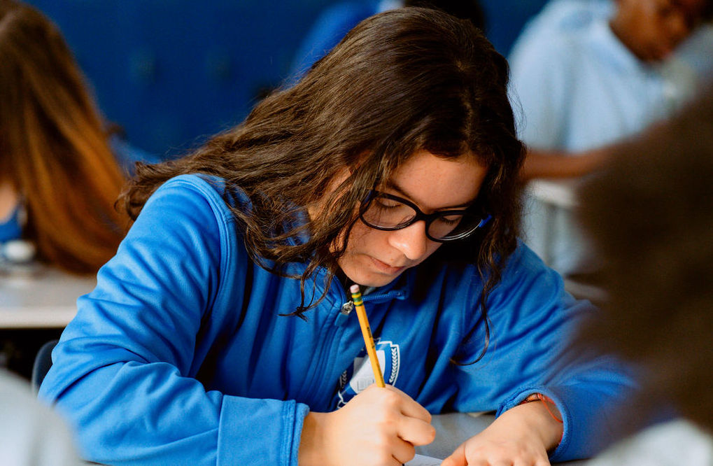 A United Charter High School Student wearing glasses and a blue sweatshirt writes with a pencil at a desk in a classroom.