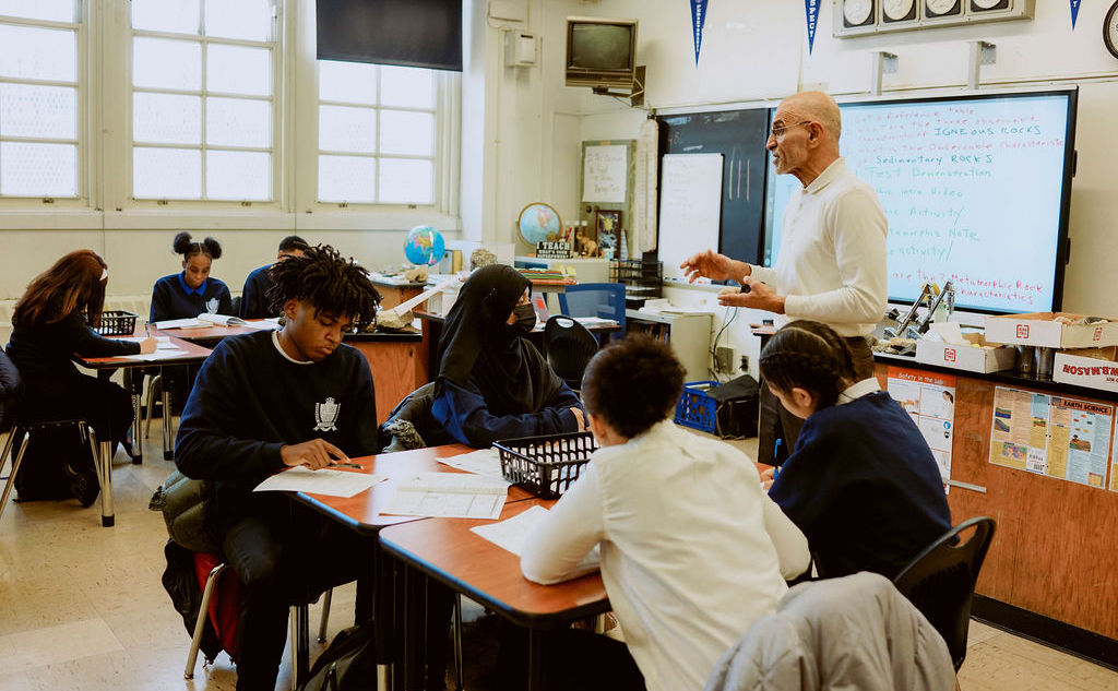 A teacher stands at the front of a classroom speaking as students sit in groups at desks, working on assignments.