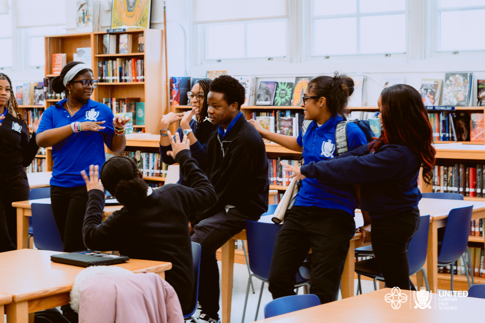 A group of students in blue uniforms stand and sit around a table in a library, talking and gesturing with their hands while discussing their upcoming HBCU Tour.
