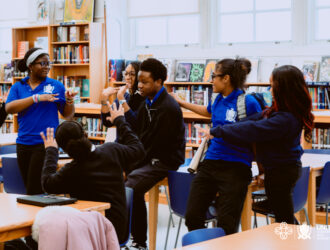 A group of students in matching blue uniforms talk and gesture energetically around tables in a school library, sharing their excitement about the upcoming HBCU Tour.