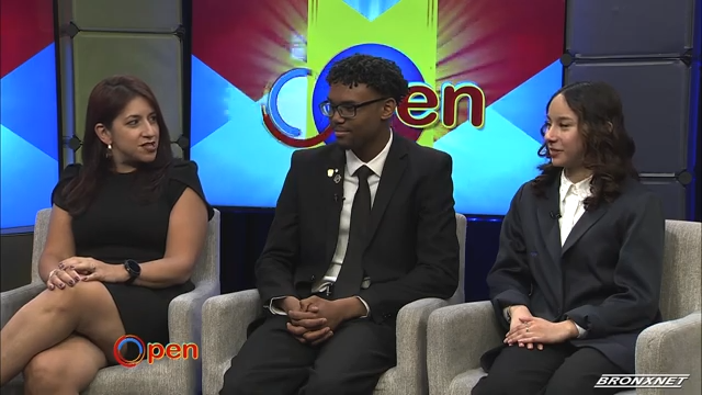 Three people in formal attire sit on gray chairs in a studio, with a colorful "Open" sign displayed on the screen behind them.