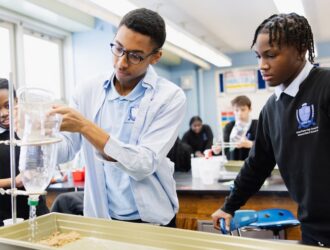 Three United Charter High School students conduct a science experiment in a classroom, using plastic bottles and trays with sand, while others work in the background.