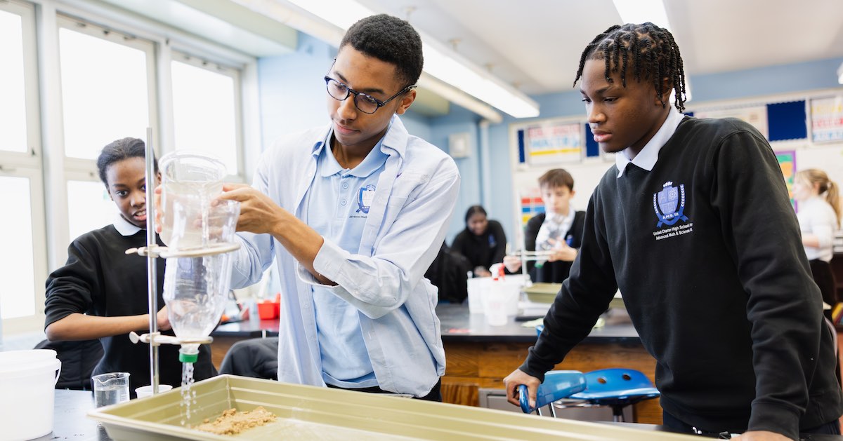 Three United Charter High School students conduct a science experiment in a classroom, using plastic bottles and trays with sand, while others work in the background.