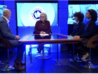 Four people sit around a glass table in a TV studio, engaged in discussion. Two men and two women face each other, with screens and blue lighting in the background.