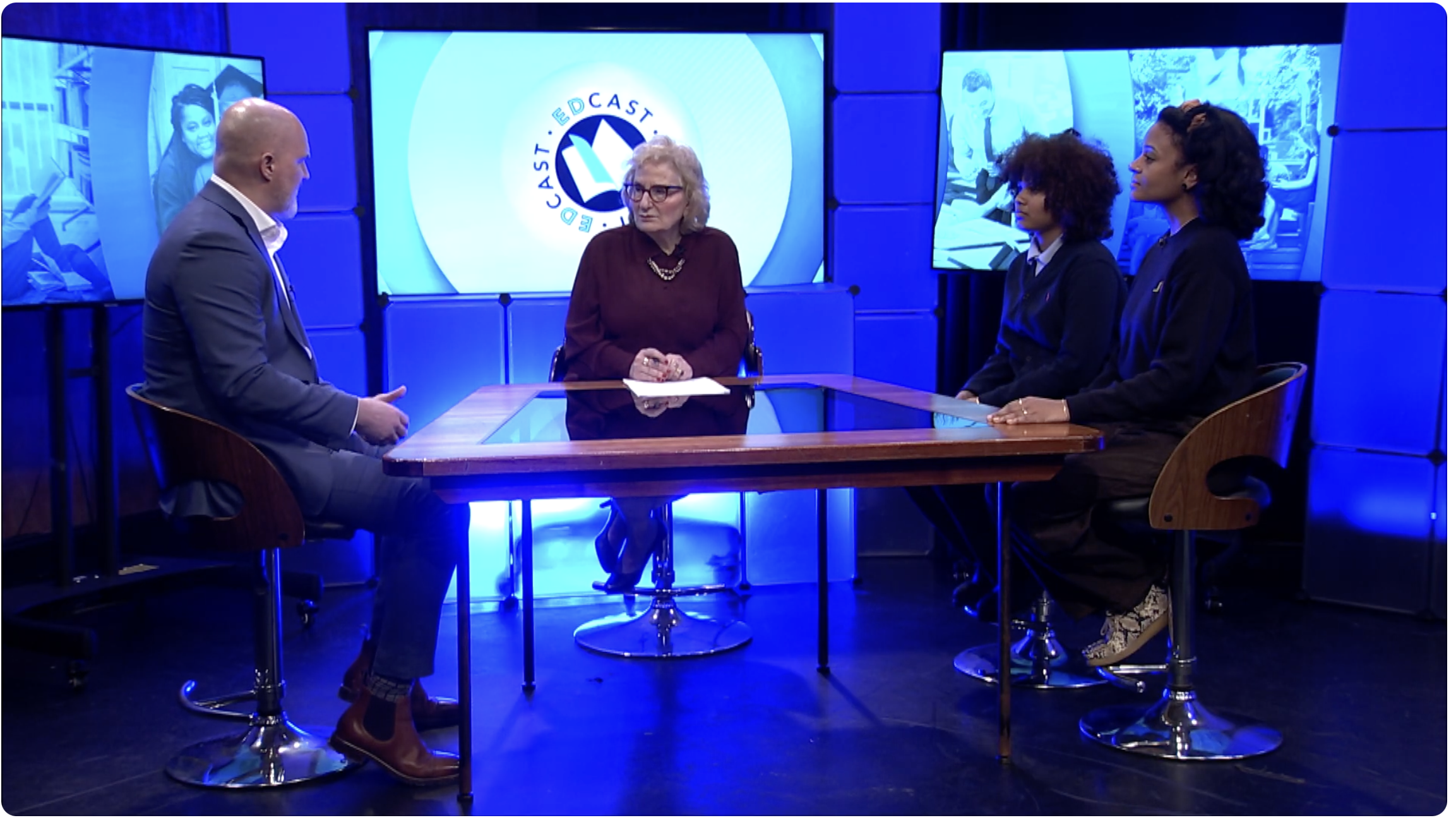 Four people sit around a glass table in a TV studio, engaged in discussion. Two men and two women face each other, with screens and blue lighting in the background.