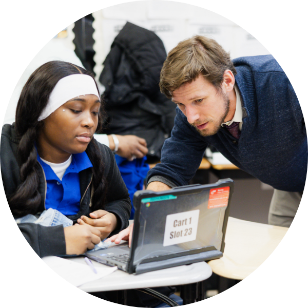 A student sits at a desk using a laptop while an instructor leans over to provide assistance in a classroom setting.