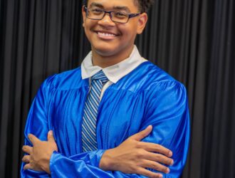 United Charter High School senior wearing a blue graduation gown and tie stands with arms crossed in front of a black curtain, smiling at the camera.