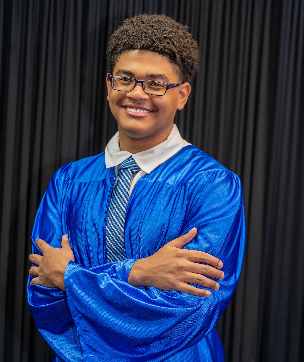 United Charter High School senior wearing a blue graduation gown and tie stands with arms crossed in front of a black curtain, smiling at the camera.