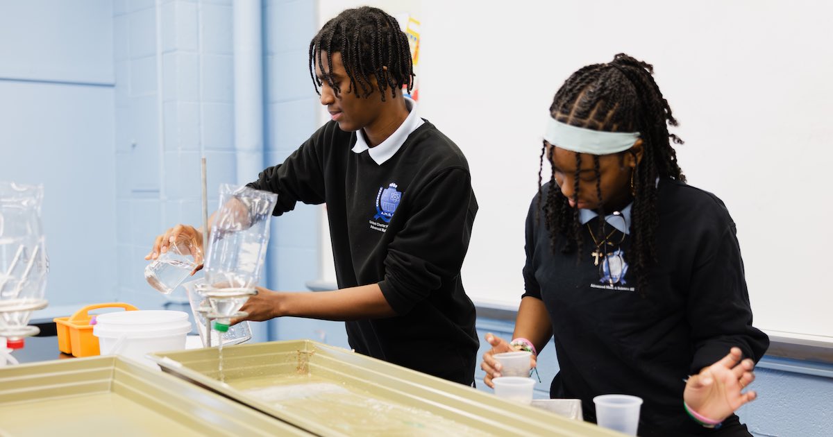 Two students from United Charter High School conduct a science experiment, pouring liquid through funnels into cups on a table with containers and lab equipment.