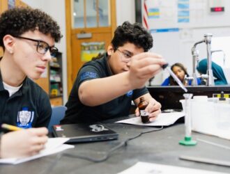 Two United Charter High School AMS II students at a lab table conduct a science experiment, with one using a dropper while the other takes notes.